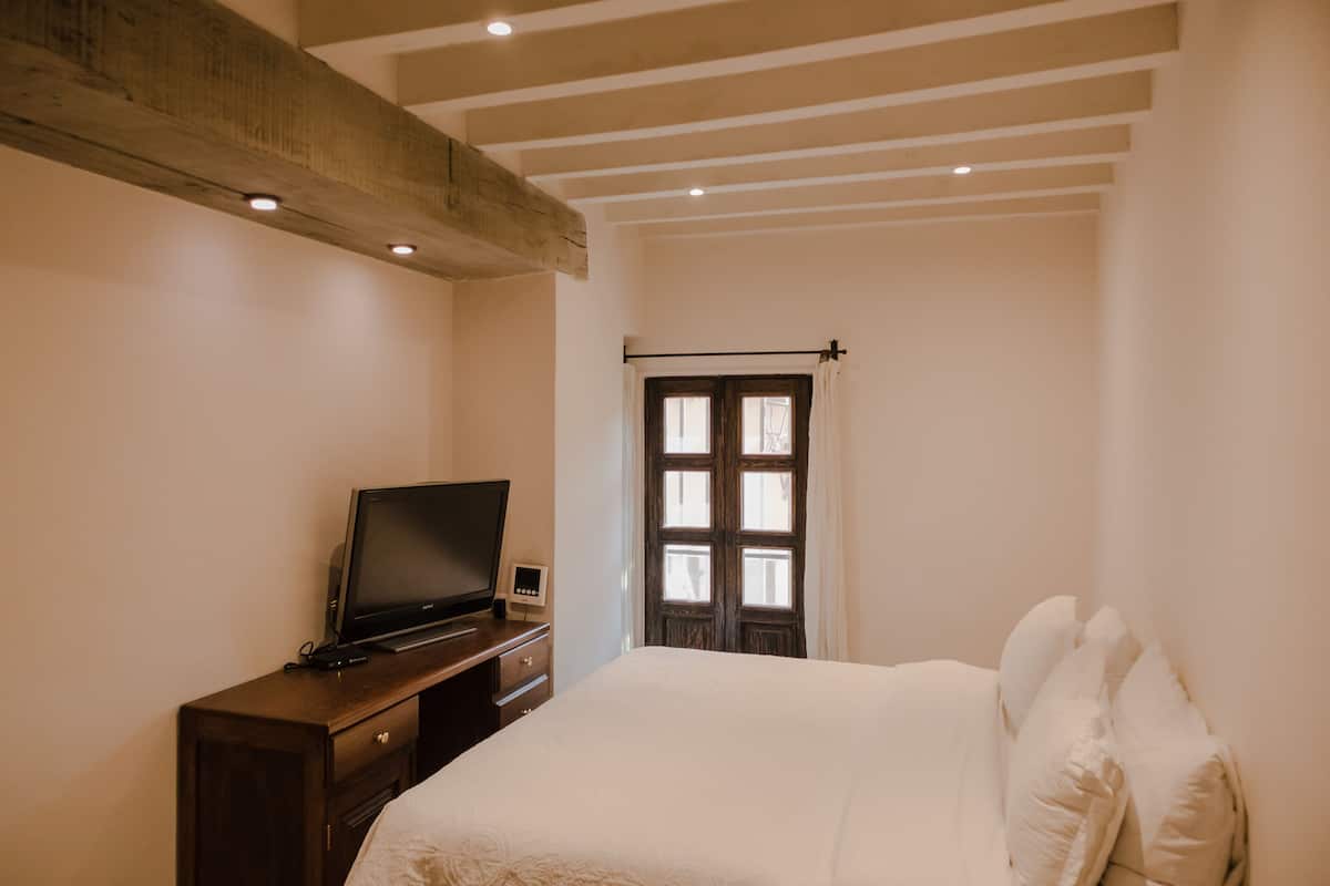 Une chambre à coucher avec un lit, un téléviseur posé sur une commode en bois et une fenêtre avec des rideaux.