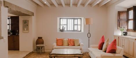 Una sala de estar con sofá, reposapiés y una mesa de centro. Se ve una ventana con cortinas, una lámpara y una zona de cocina al fondo.