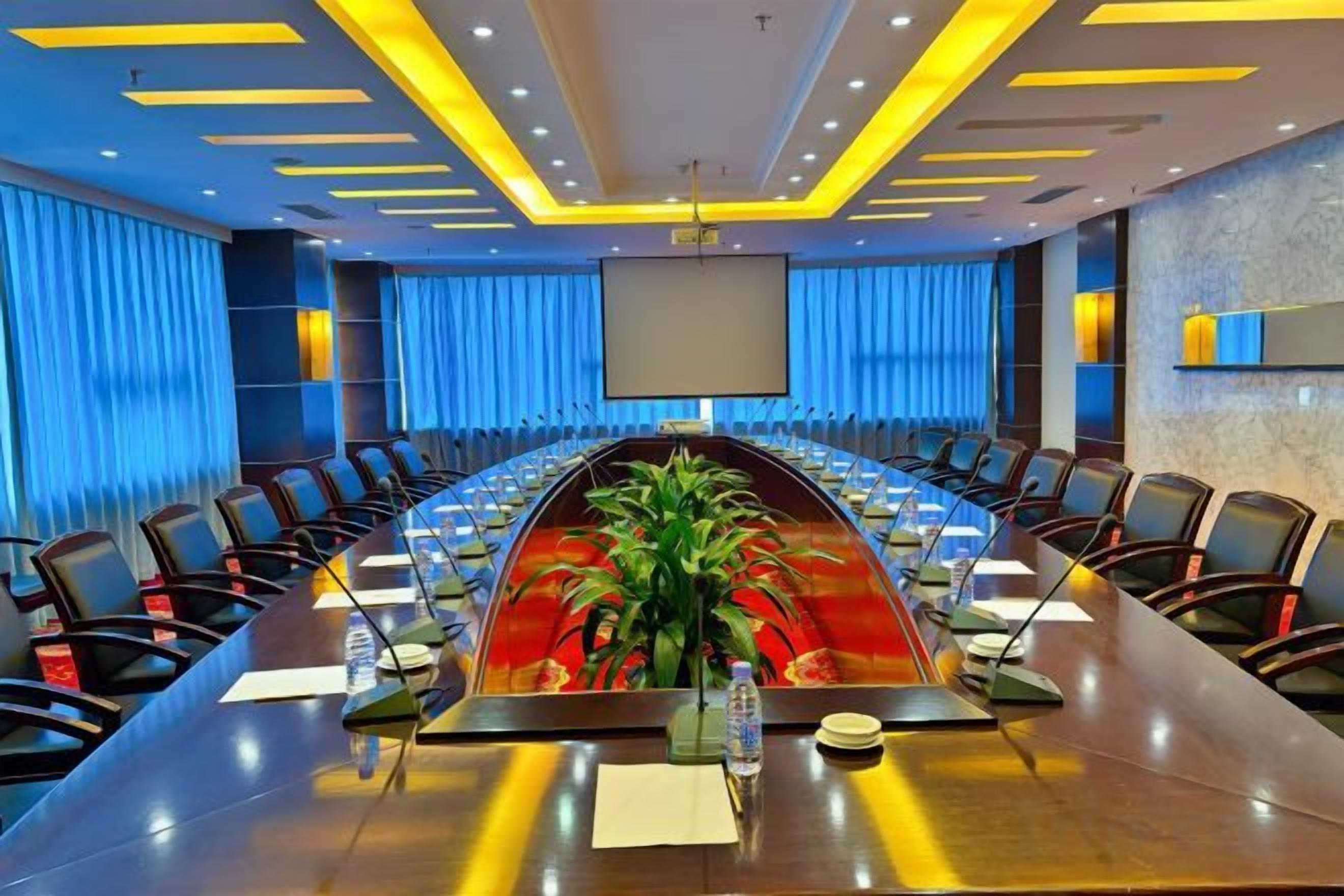 Un salón de conferencias con una larga mesa, sillas, una pantalla de proyector y una planta.