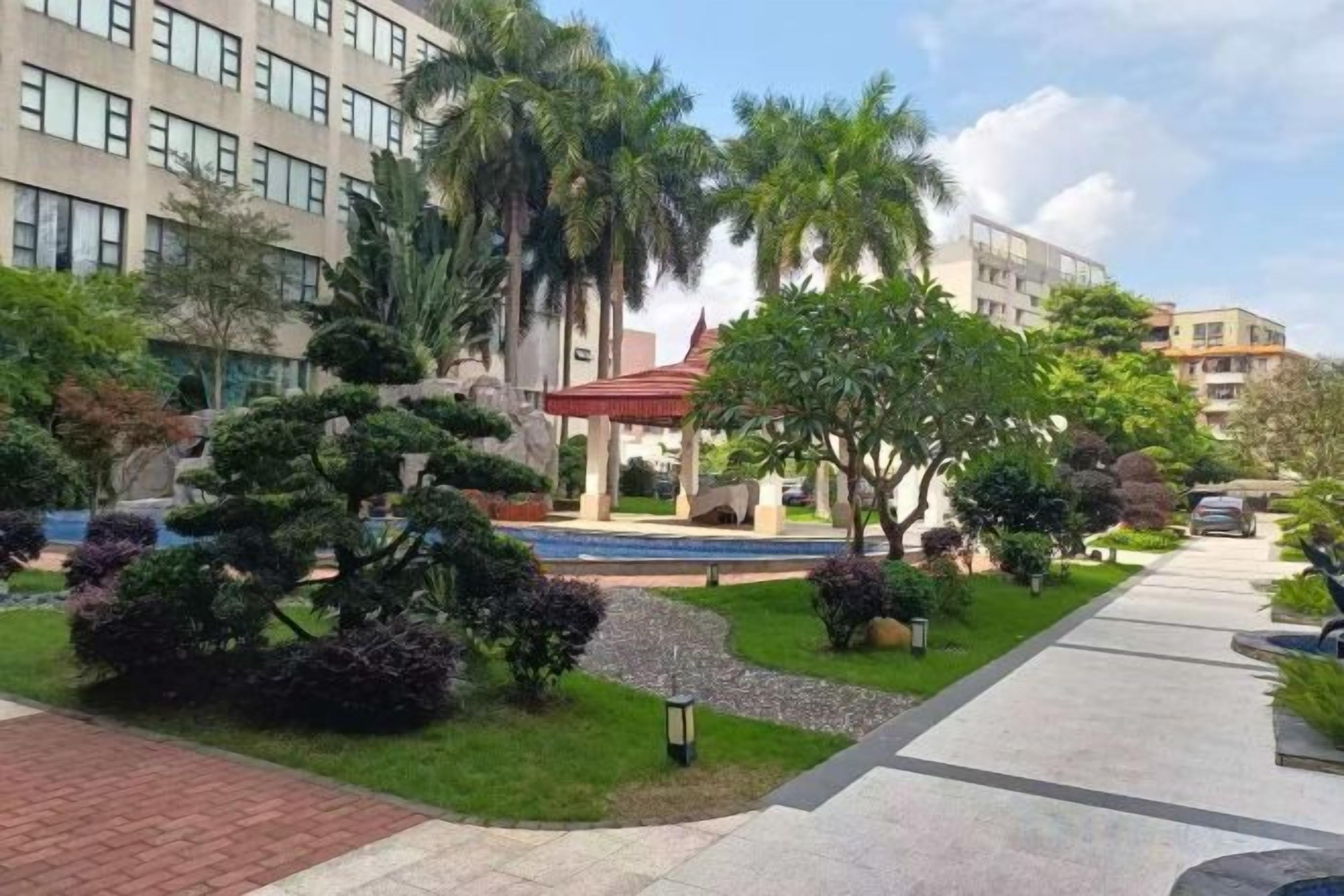 A landscaped courtyard with a paved walkway, palm trees, and a building with a red roof in the background.