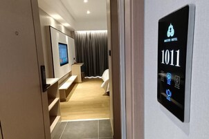 Quarto de hotel moderno com tela digital exibindo o número do quarto 1011, televisão, cama e banheiro com chuveiro.