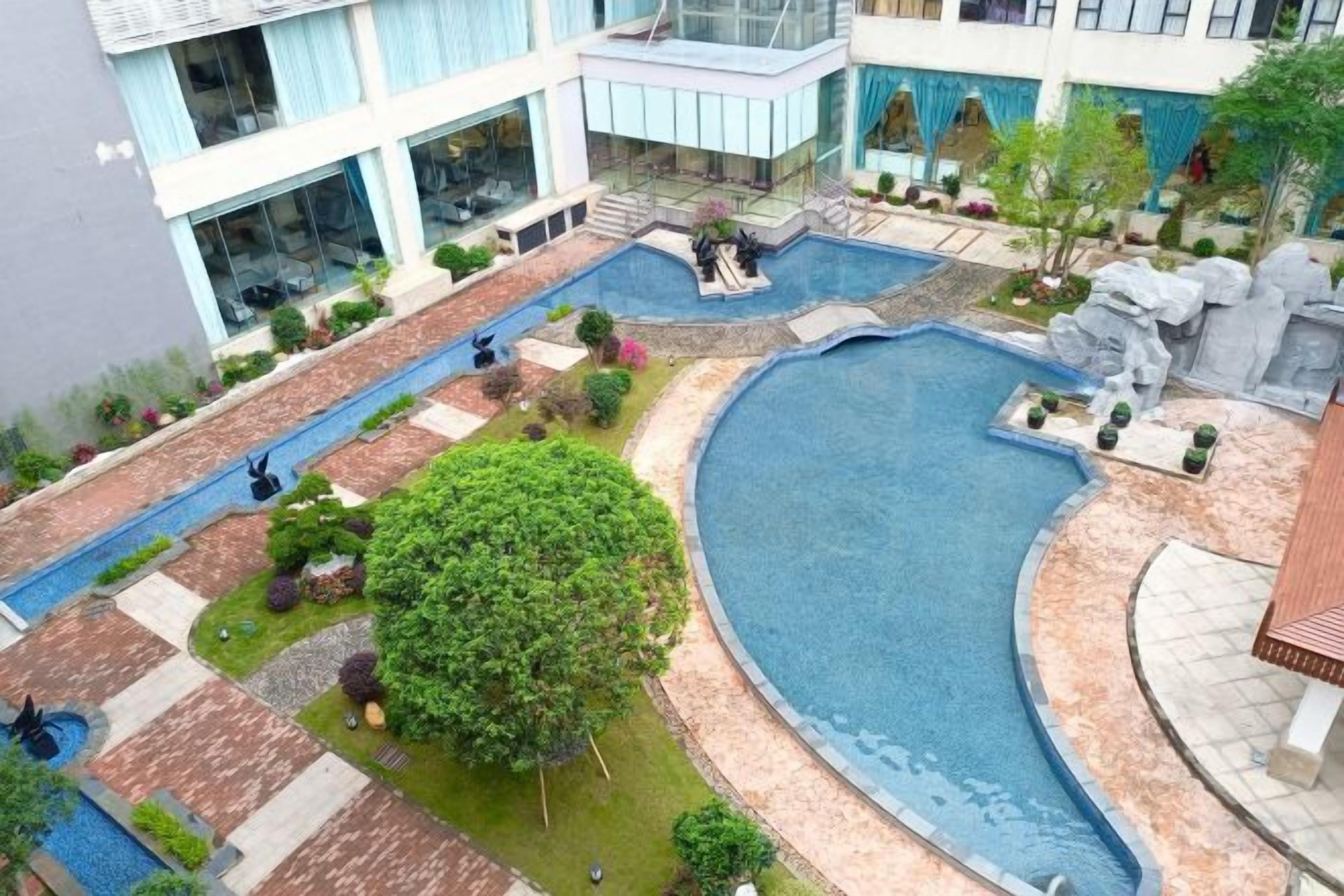 Un espace piscine comprenant plusieurs bassins, entouré de verdure et bordé d’un bâtiment doté de grandes fenêtres.