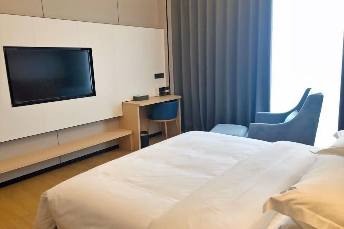 A hotel room with a bed, a desk, a chair, a TV mounted on the wall, and a window with curtains.