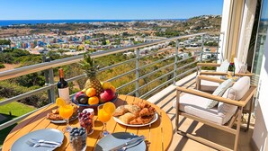 A balcony with a table set for breakfast, including croissants, fruit, and juice, overlooking a coastal town.