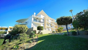 A modern multi-story hotel with balconies, surrounded by well-maintained gardens and a clear blue sky.