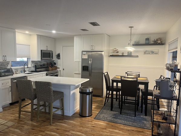A modern kitchen with white cabinets, a stainless steel refrigerator, a microwave, and a built-in oven. There is a dining area with a table and chairs, and a trash can near the kitchen.
