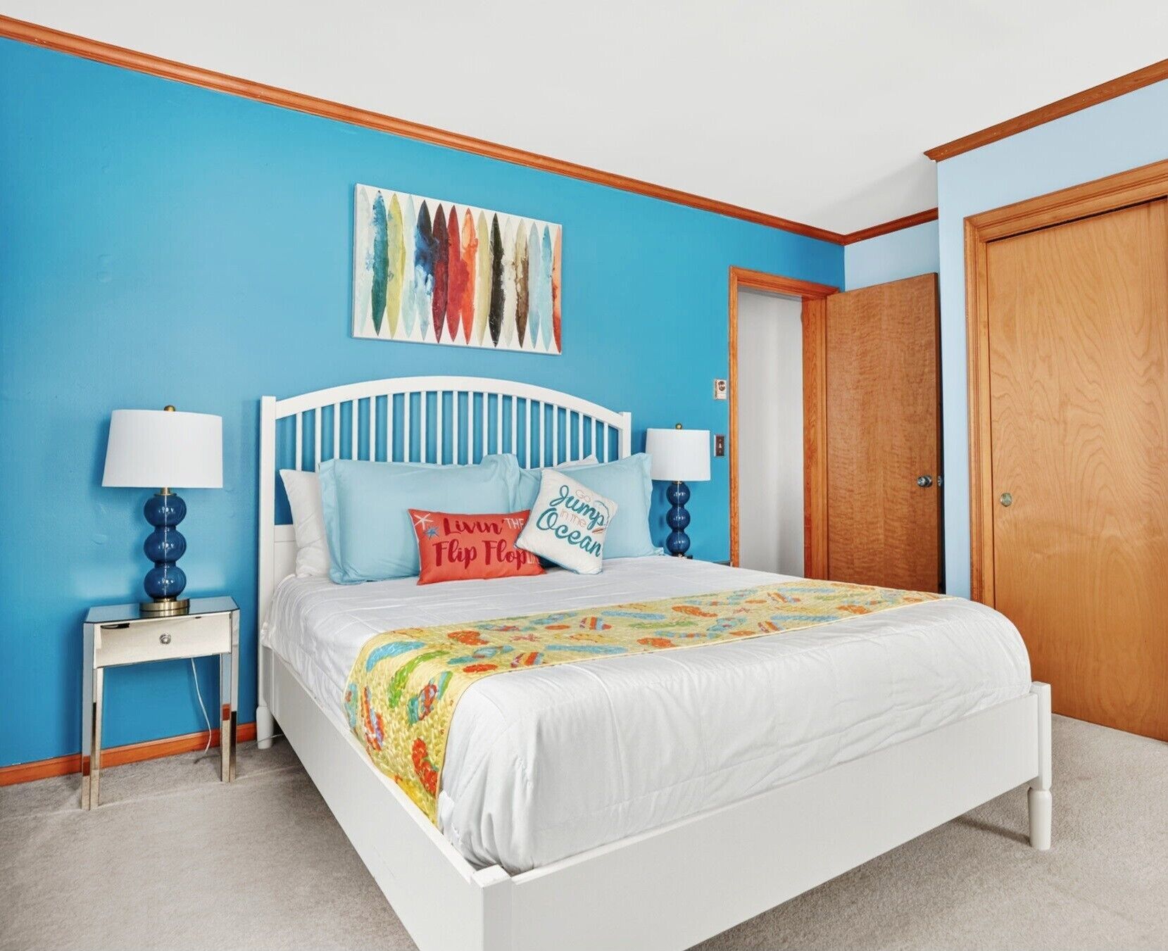 A bedroom with a white bed, blue walls, a wooden door, and a colorful artwork above the bed.