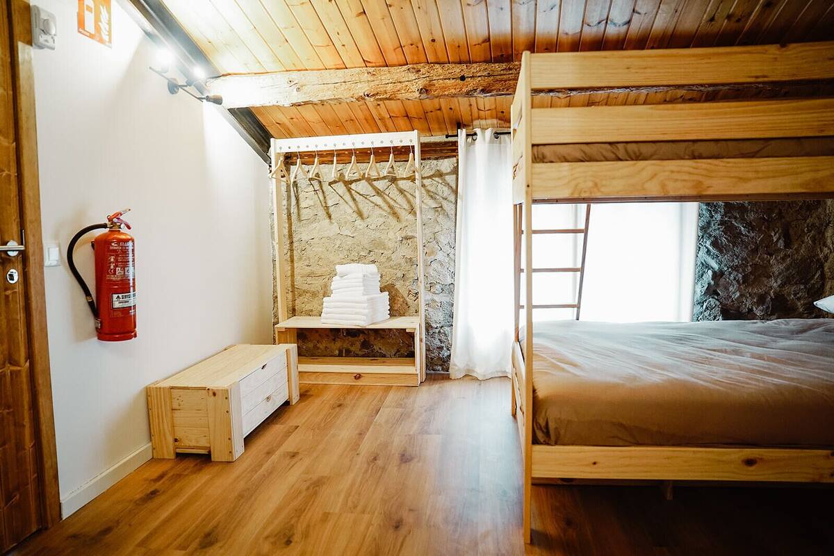 A bunk bed room with a wooden ceiling, a fire extinguisher, a stack of towels, and a window with a view of a rocky exterior.
