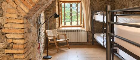 A rustic room with exposed brick walls, a sloped ceiling, bunk beds, a window with curtains, a desk with a chair, and a floor lamp.