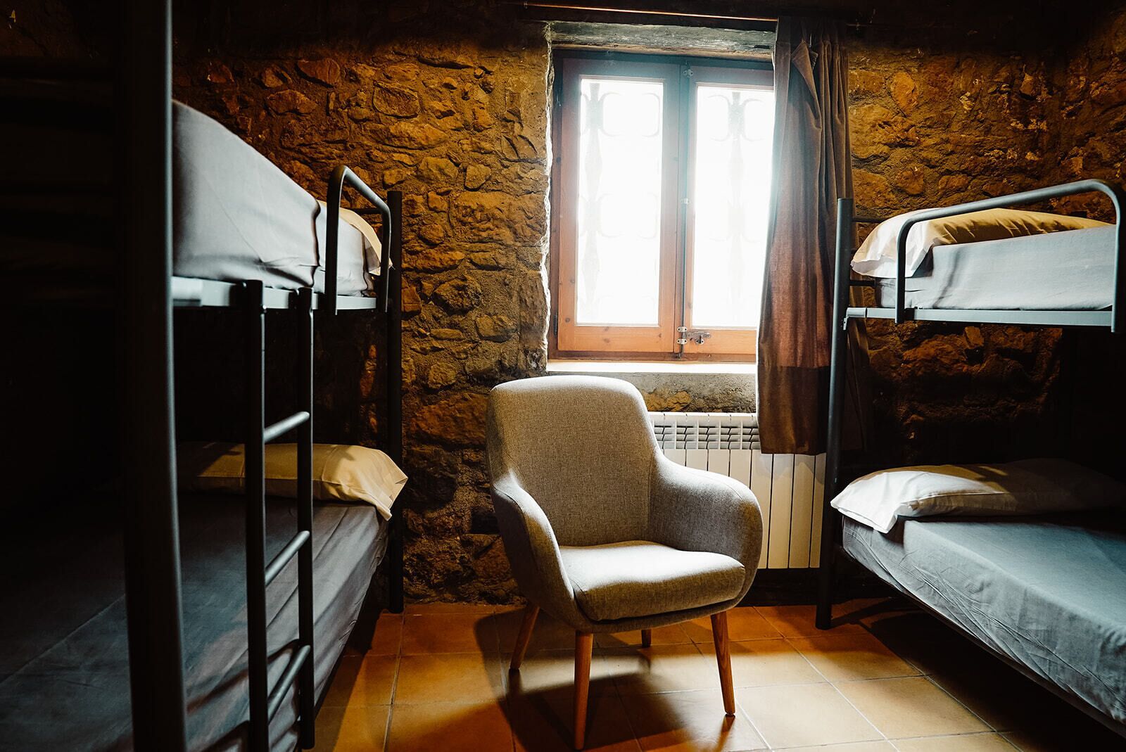 A dormitory room with bunk beds, a chair, a window with curtains, and a radiator.