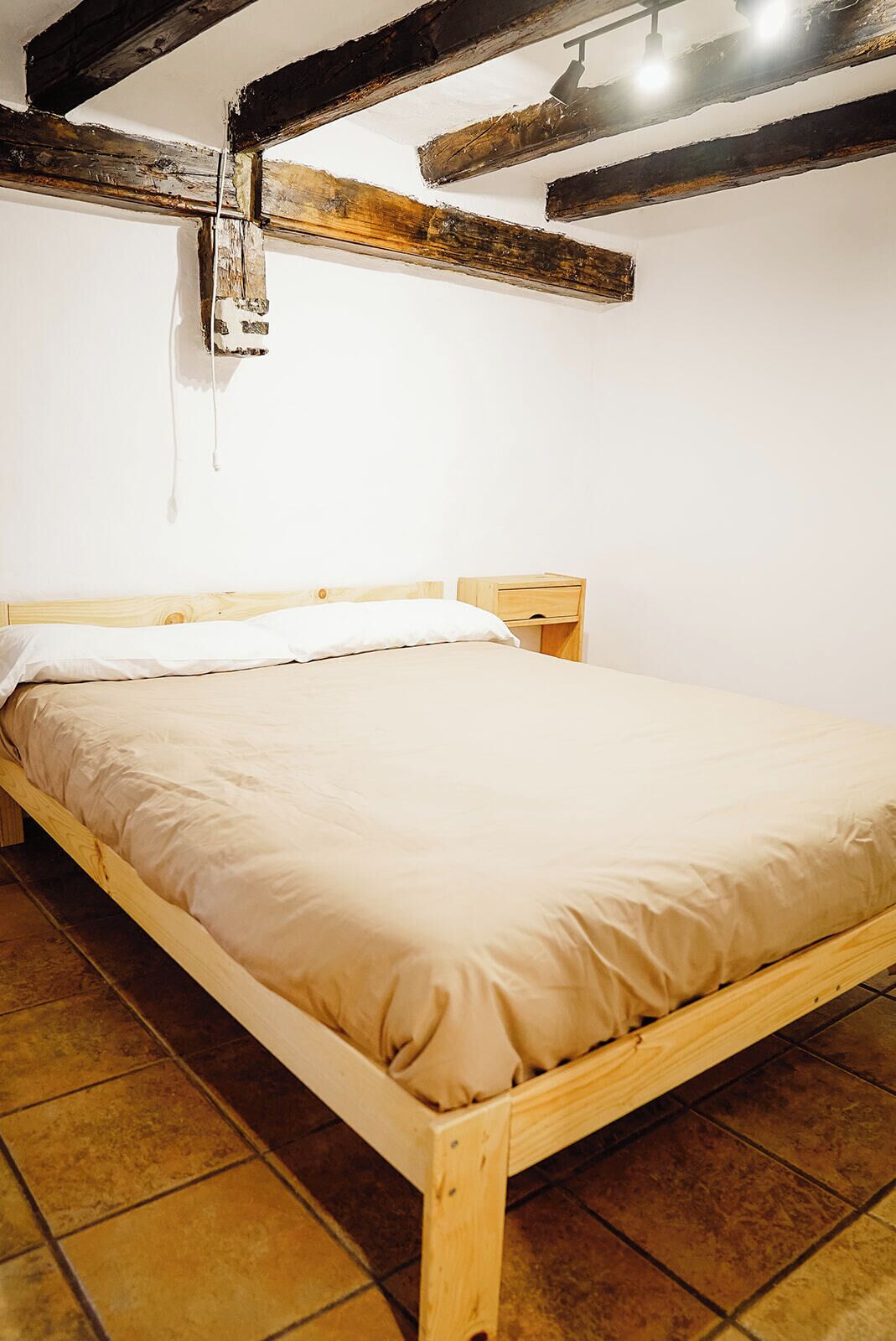 A single bed with a wooden frame and a beige mattress in a room with exposed wooden beams and tiled flooring.