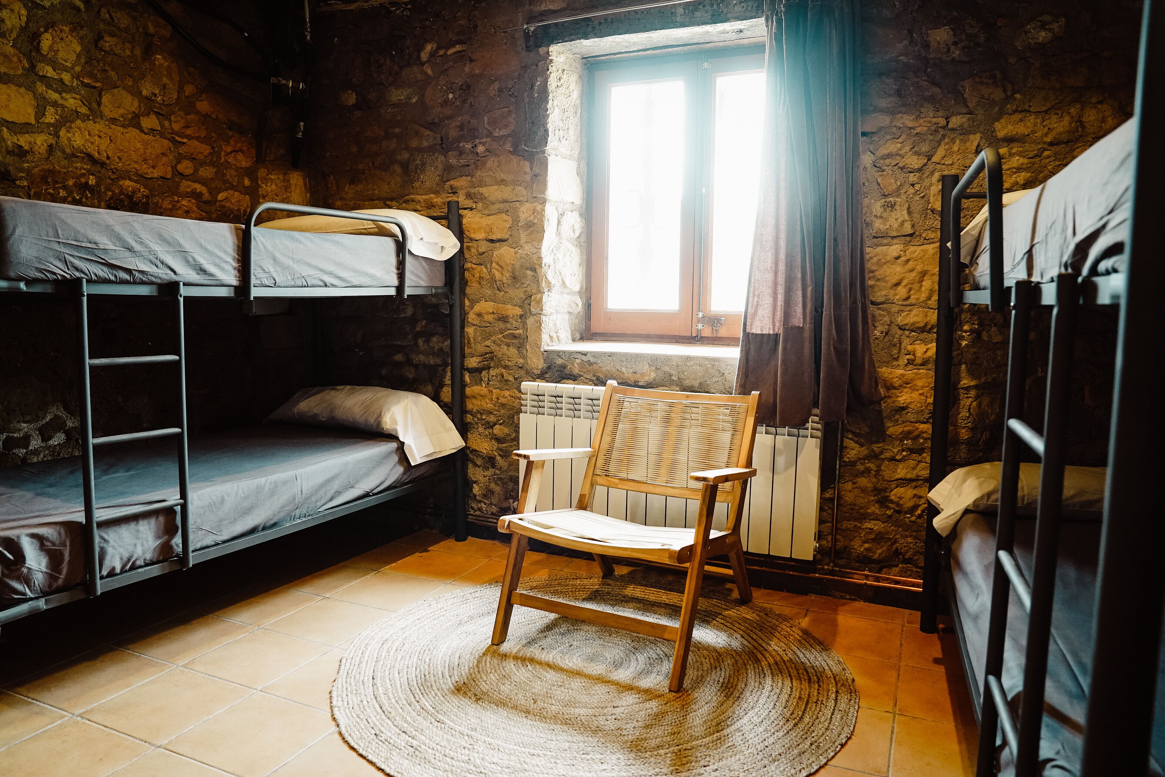 A dormitory room with bunk beds, a wooden chair, a window with curtains, and a stone wall.