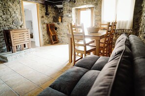A cozy living room with a sofa, wooden dining table, chairs, and a stone wall. - Cases Altes de Posada (Navès)