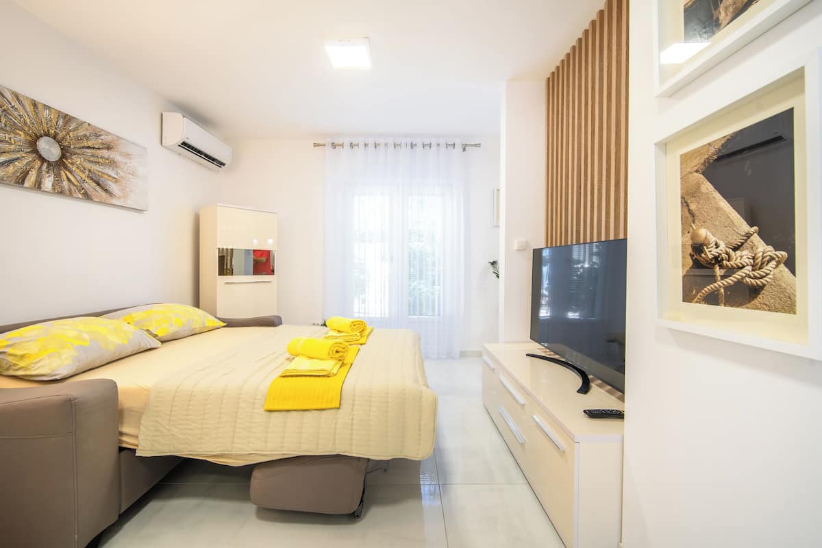 A hotel room with a bed, a flat-screen TV, a desk, and a window with sheer curtains.
