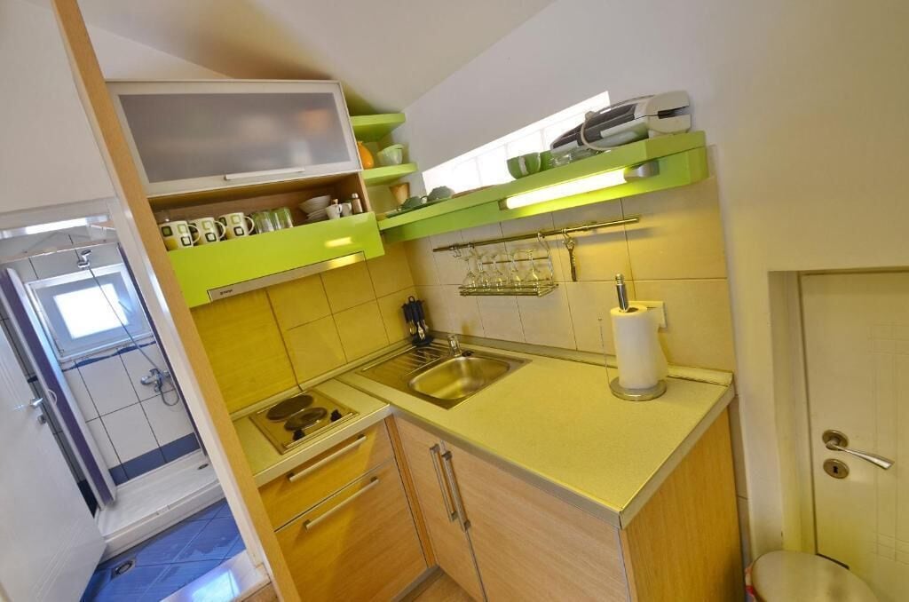 A compact kitchen with a sink, a stove, and a countertop. There are shelves above the counter holding dishes and a small refrigerator.