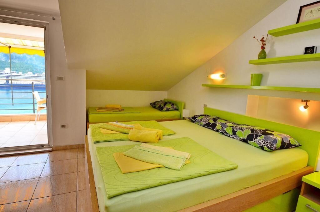 A room with two beds, a balcony with a view of the sea, and a green shelf with decorative items.