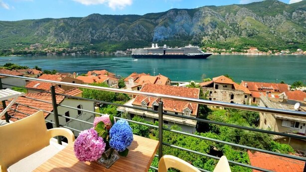 A balcony with a table, chairs, and flowers, overlooking a body of water with a cruise ship and mountainous landscape in the background. - Palma Apartments (Kotor)