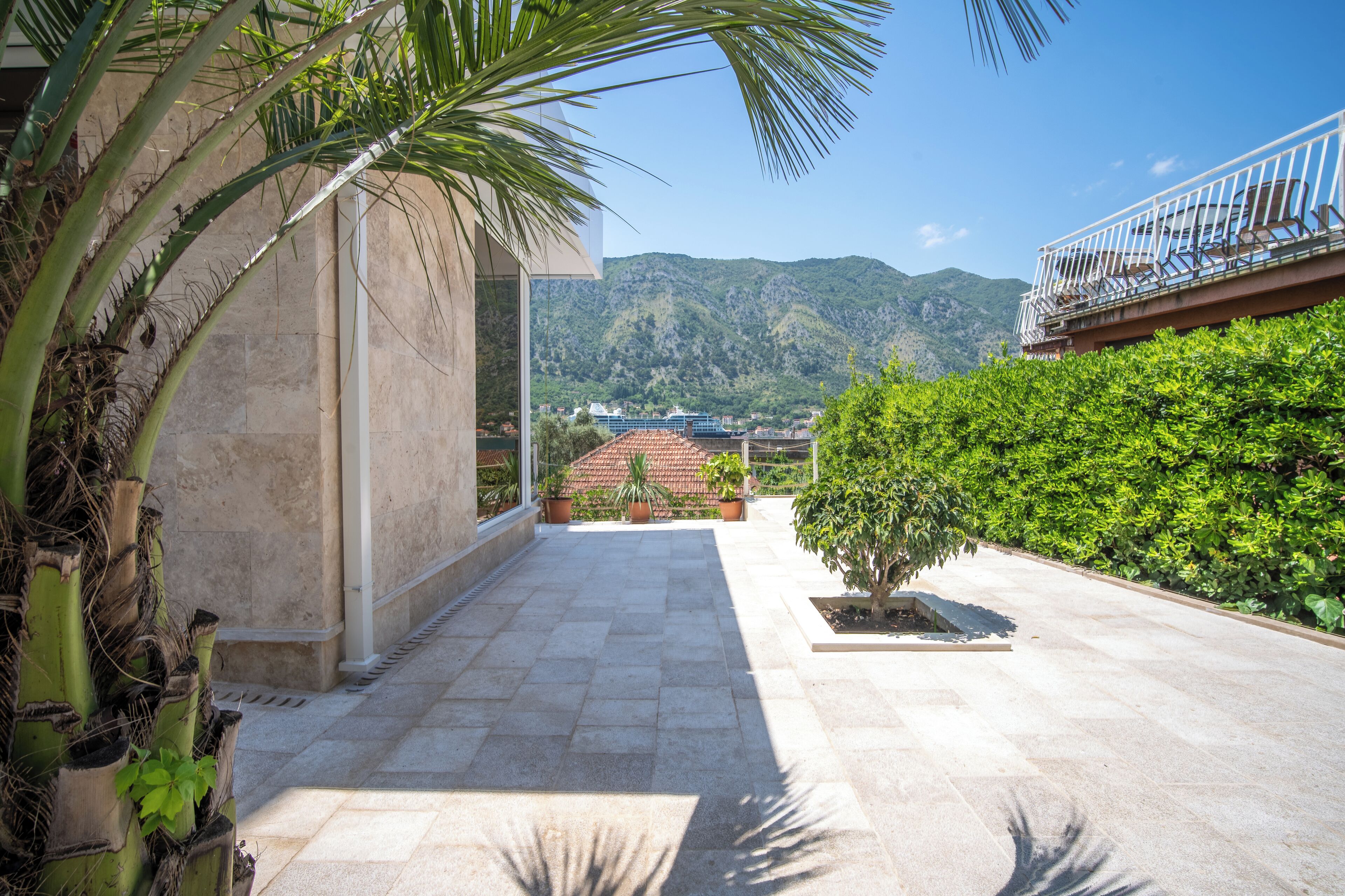 A paved outdoor area with a view of mountains, a balcony with a railing, and potted plants.