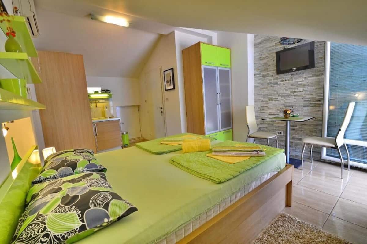 A bedroom with a bed, a desk, a chair, a TV, and a kitchenette.