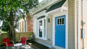 Une petite maison avec une porte bleue, une cheminée en briques et une petite terrasse avec deux chaises rouges et une table.