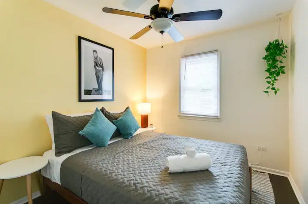 Una cama bien hecha con colcha gris, un ventilador de techo, un cuadro enmarcado en la pared, una lámpara de noche y una ventana con persianas.