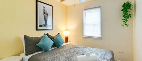 Una cama bien hecha con colcha gris, un ventilador de techo, un cuadro enmarcado en la pared, una lámpara de noche y una ventana con persianas.