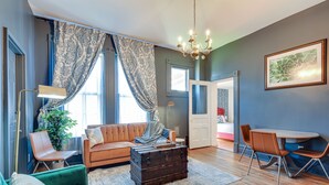 A living room with a green sofa, a brown leather chair, a wooden table with two chairs, a large window with blue curtains, and a chandelier. - Experience History in Style in an 8BR/8BA Private Guesthouse! (Columbus)