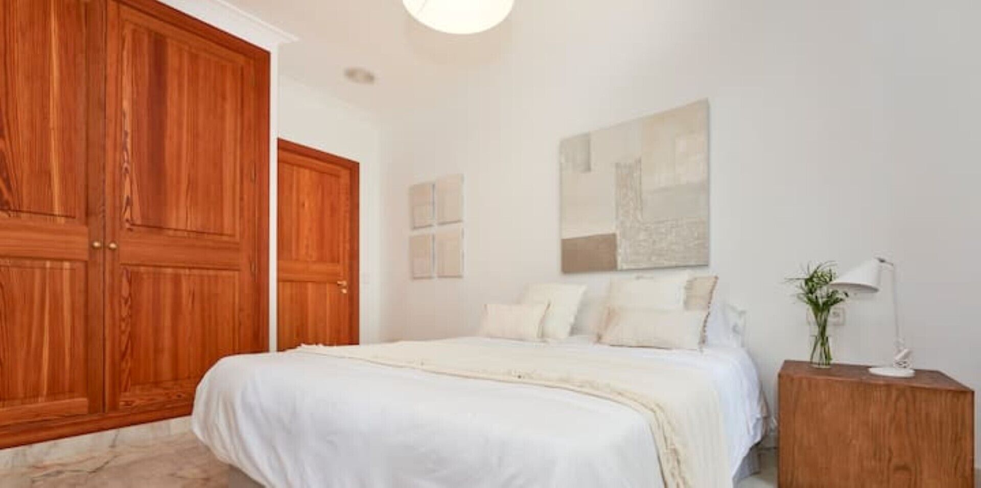 A bedroom with a large wooden wardrobe, a bed with white bedding, a bedside table with a lamp, and a wall-mounted artwork.
