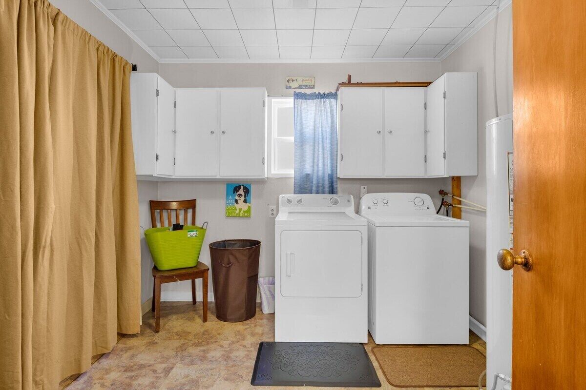 A laundry room with two white washing machines, a wooden chair, a green basket, and a trash can.