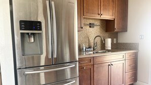A stainless steel refrigerator with a water dispenser, wooden cabinets, a sink, and a patterned rug in a kitchen. - Bohemian Bungalow on Byron Road (Fort Myers)
