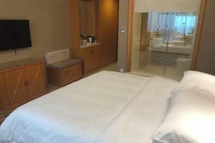A hotel room with a bed, a television, a wooden wardrobe, and a bathroom visible through a glass door.