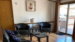 A room with a wooden floor, a black sofa with a patterned pillow, two black armchairs, a small table with a lamp, and a framed picture on the wall. - House 70 Oasis (Put-in-Bay)