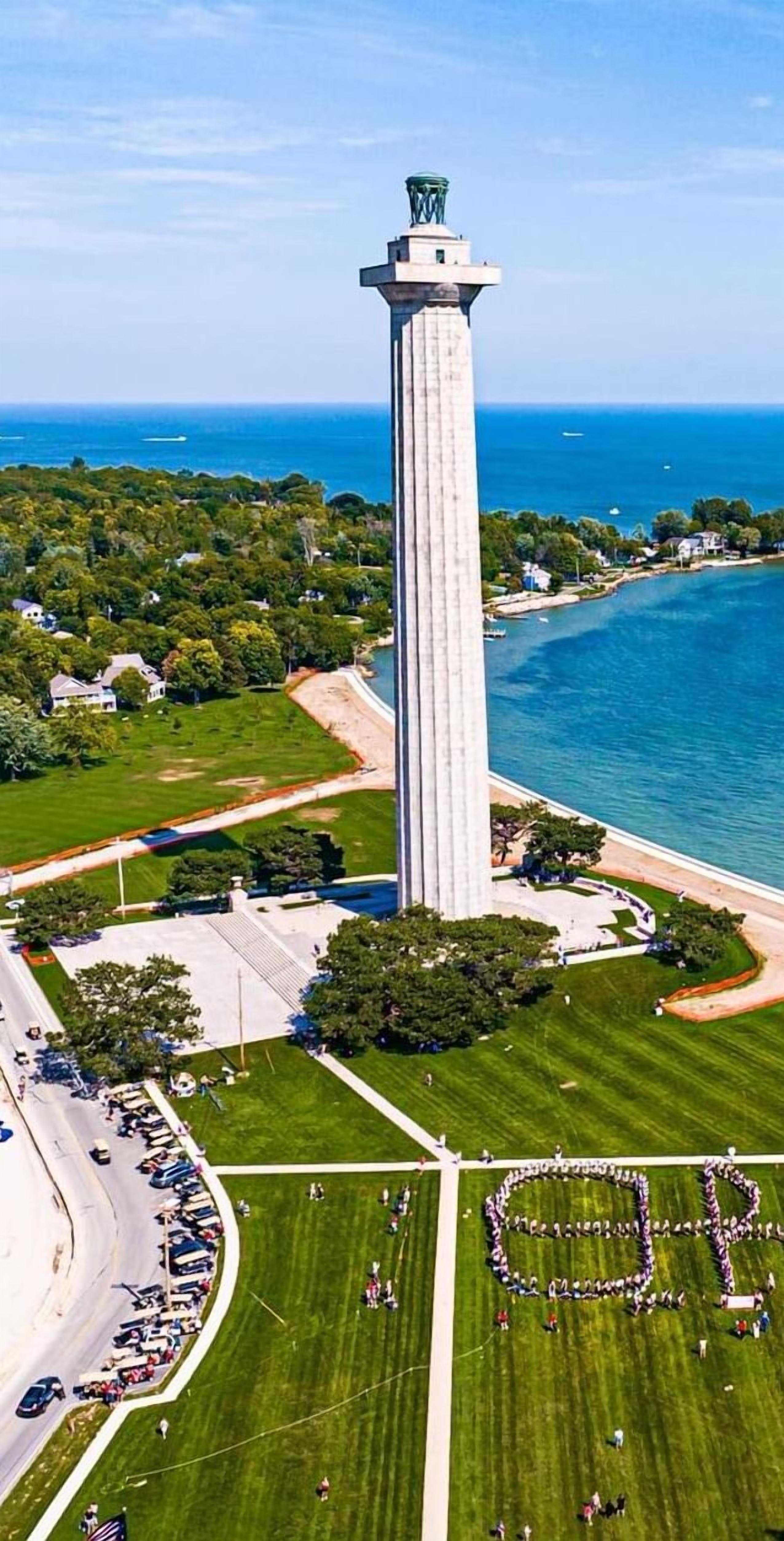 Vista aérea de un monumento alto con base circular, rodeado por un parque y una masa de agua.