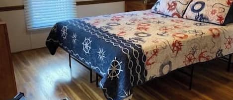 A bedroom with a wooden floor, a bed with a patterned bedspread, a nightstand with a lamp, and a window with blinds.