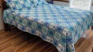 A bedroom with a wooden bed, a nautical-themed bedspread, a nightstand with a lamp, and a window with blinds. - House 70 Oasis (Put-in-Bay)