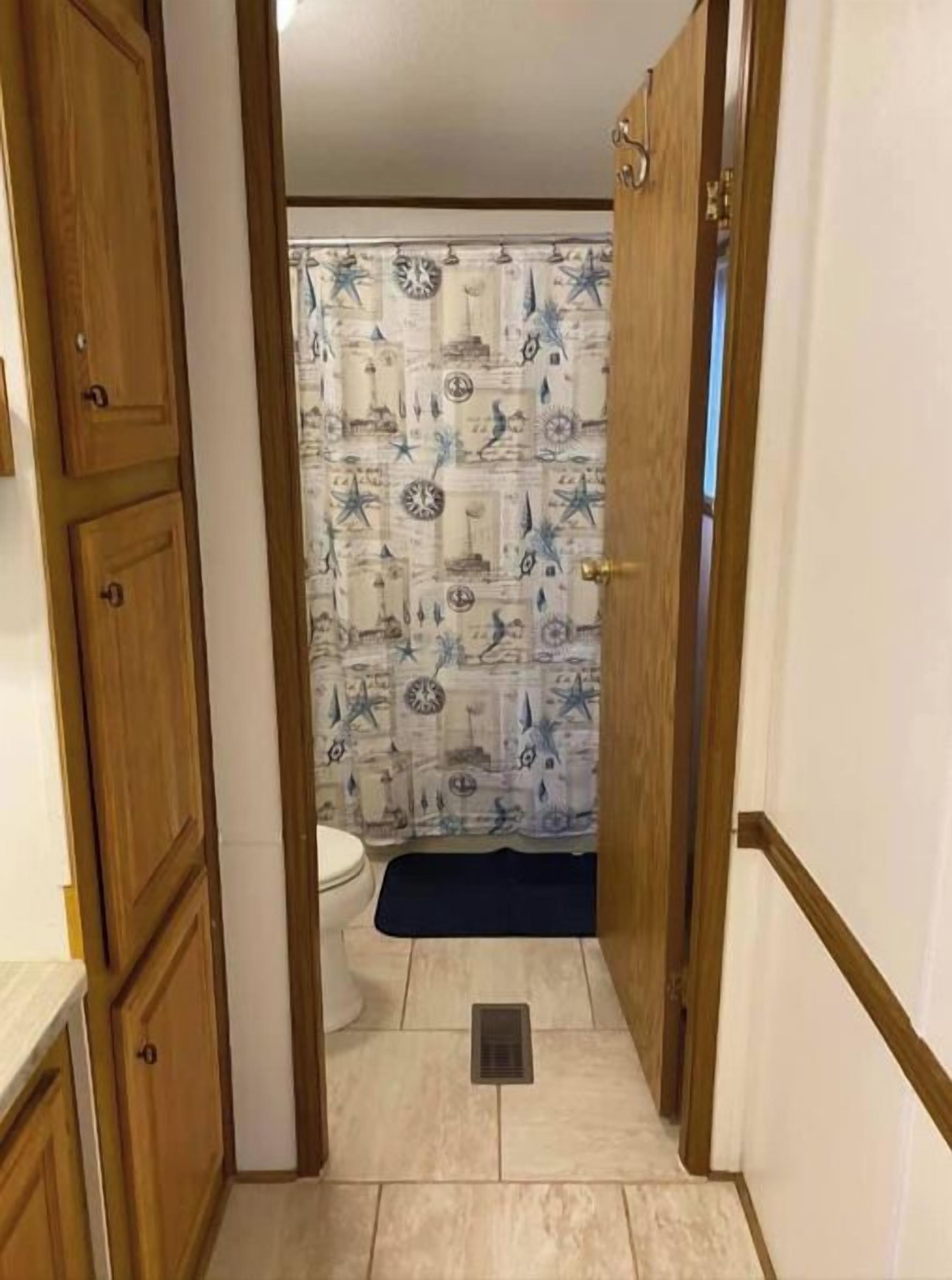 A narrow hallway leading to a bathroom with a patterned shower curtain, wooden cabinets, and a tiled floor.
