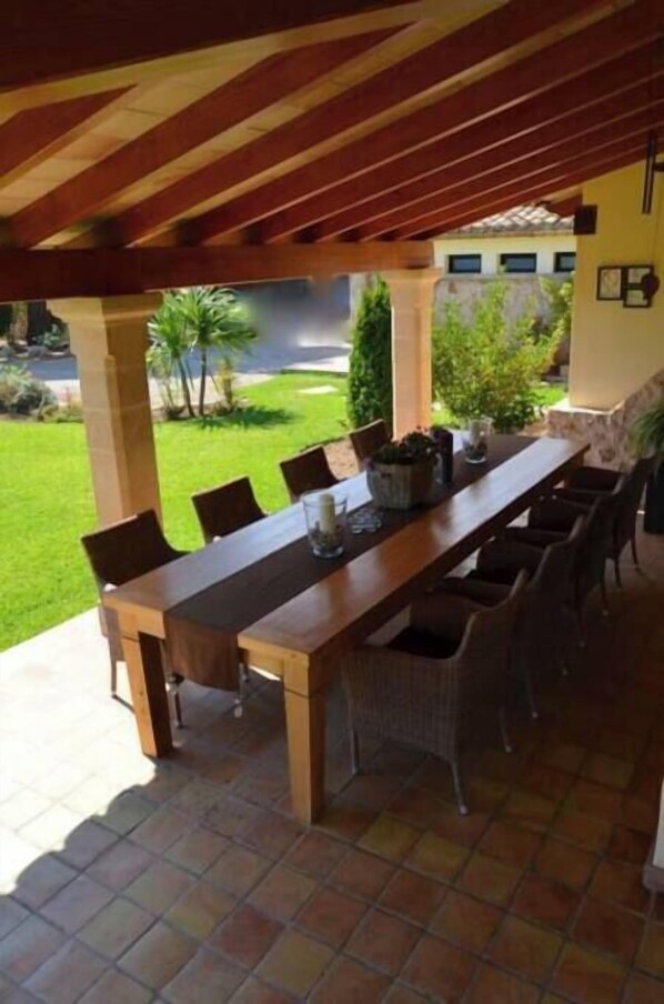 An outdoor dining area with a long wooden table and wicker chairs, under a wooden pergola with a tiled floor.