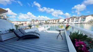 A wooden deck with lounge chairs and a small table, overlooking a marina with boats and buildings.