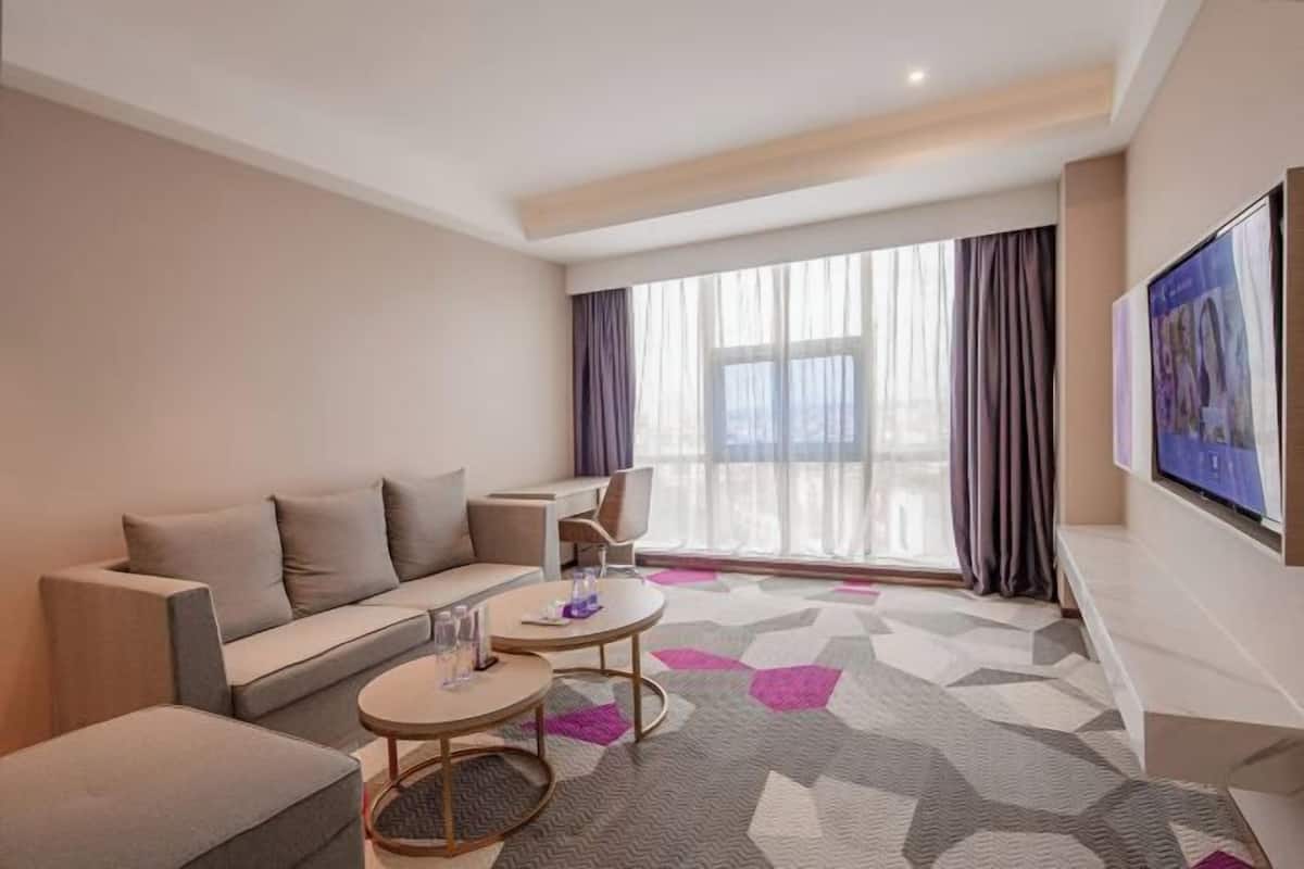 A modern hotel room with a sofa, two round tables, a flat-screen TV, and a large window with curtains.
