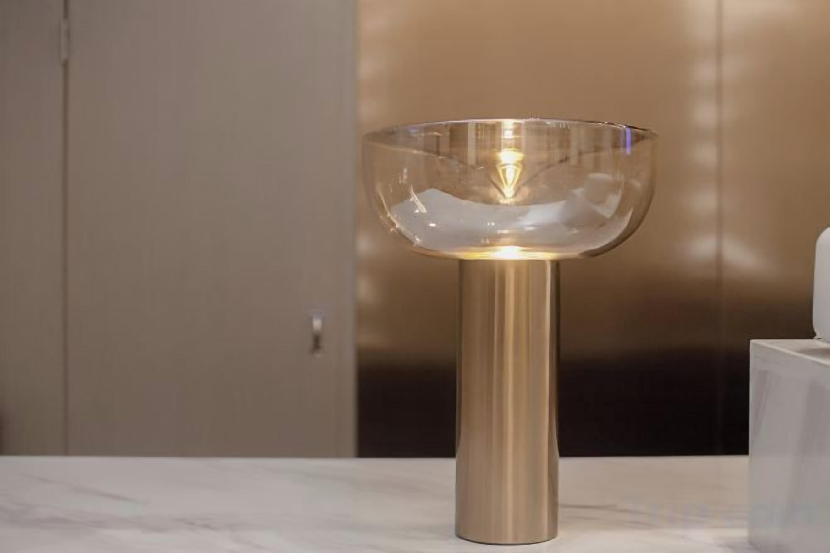 Luminária de mesa com cúpula de vidro transparente e base metálica sobre uma superfície de mármore.