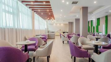 A modern hotel lobby with rows of seating, a reception area, and a view of the kitchen.