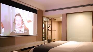 Quarto de hotel com uma cama, uma tela de TV exibindo uma pessoa e uma porta deslizante para um banheiro.