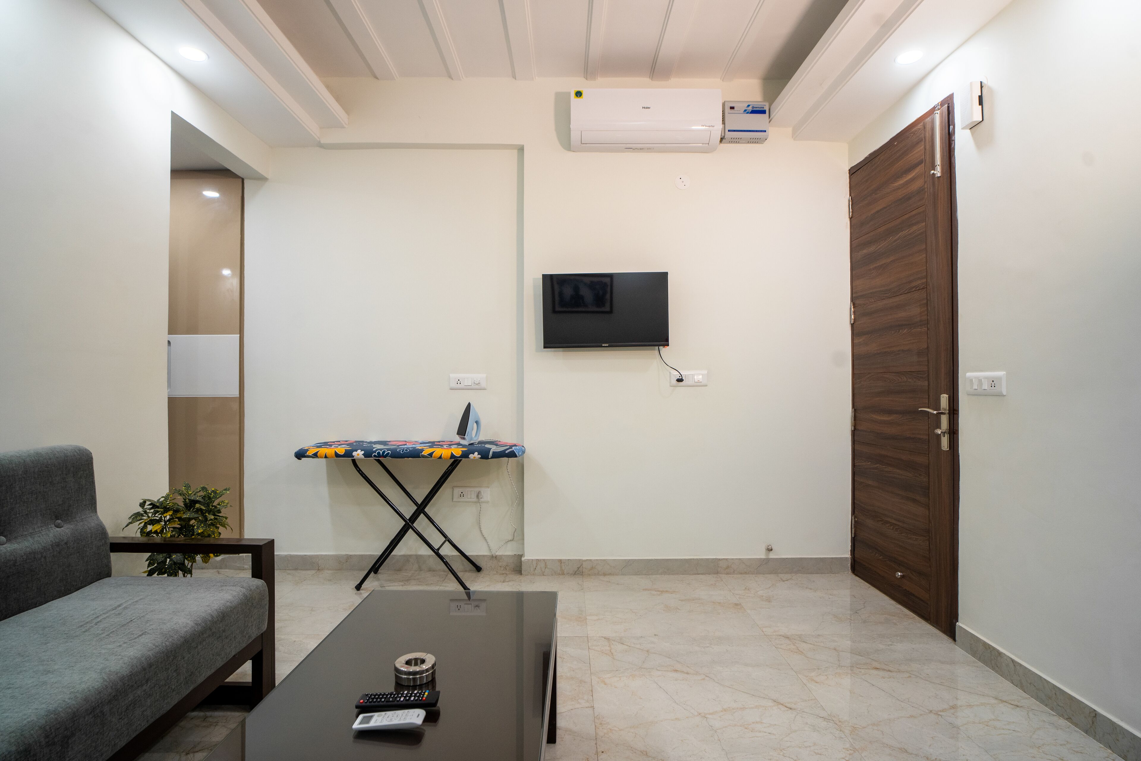 A living room with a sofa, a coffee table with remote controls, an ironing board, a television mounted on the wall, and a wooden door.
