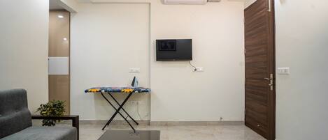 Una sala de estar con un sofá, una mesa de centro con controles remotos, una tabla de planchar, un televisor montado en la pared y una puerta de madera.