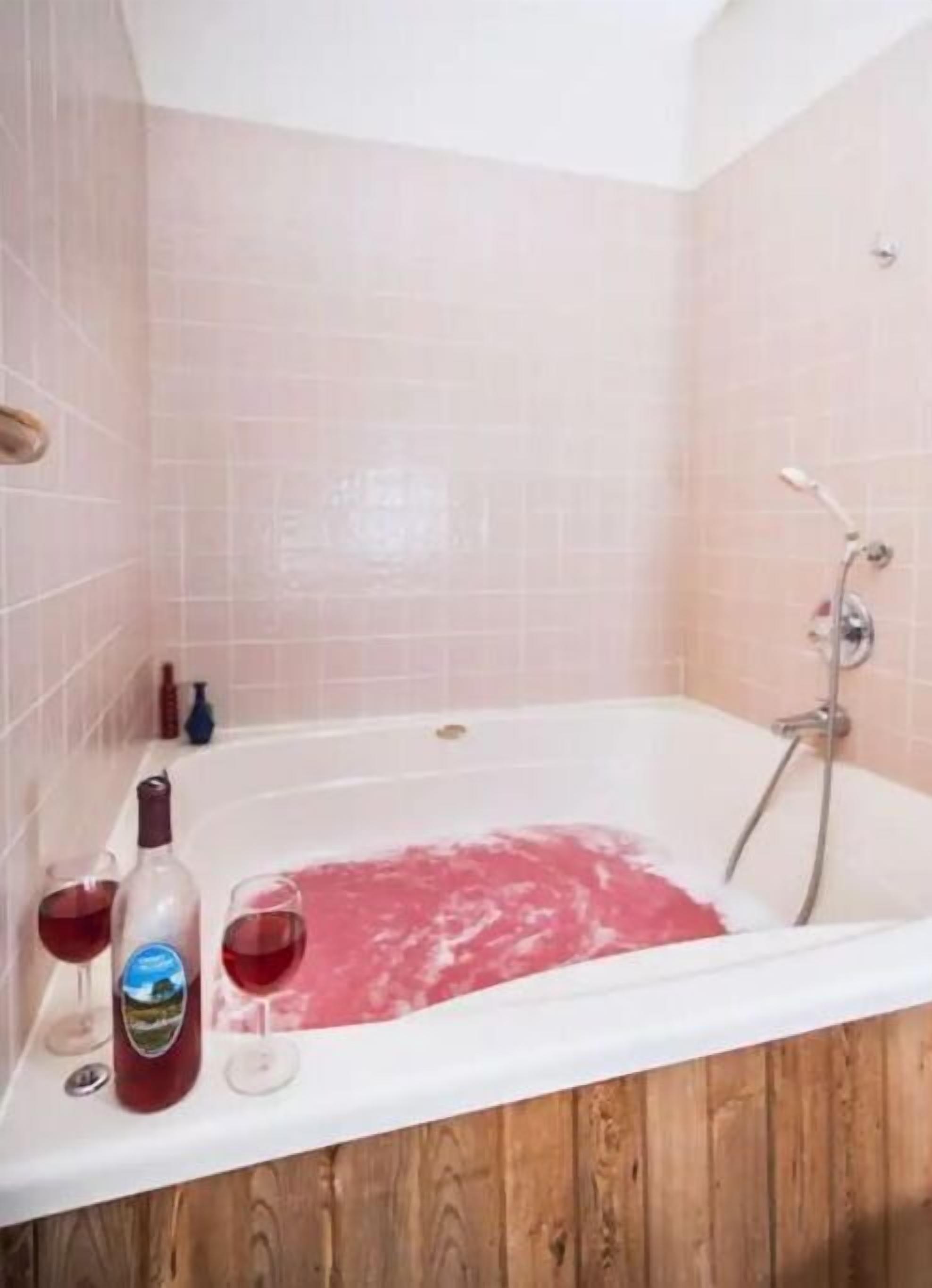 Un baño con una bañera llena de agua rosa, un cabezal de ducha y dos botellas de líquido rojo al lado.