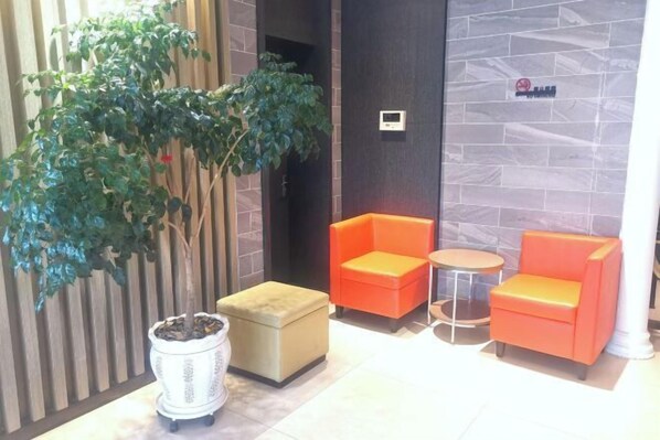 Un hall d’entrée avec une plante en pot, deux fauteuils orange, une petite table et une poubelle blanche.
