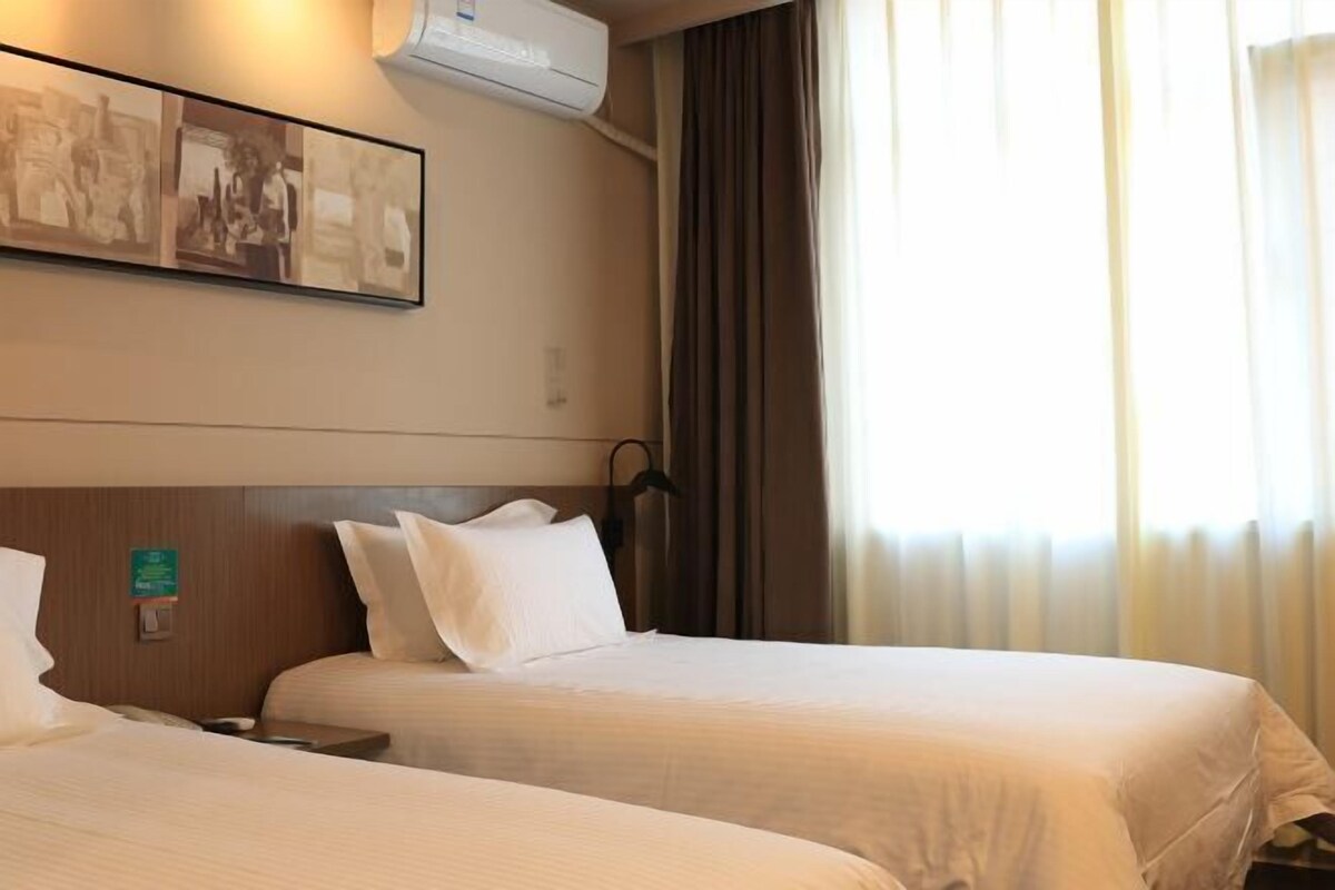 A hotel room with two beds, a wall-mounted air conditioner, a framed picture, and a window with curtains.