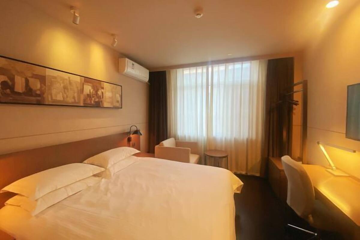 A hotel room with a bed, a desk, a chair, a small table, a lamp, and a window with curtains.
