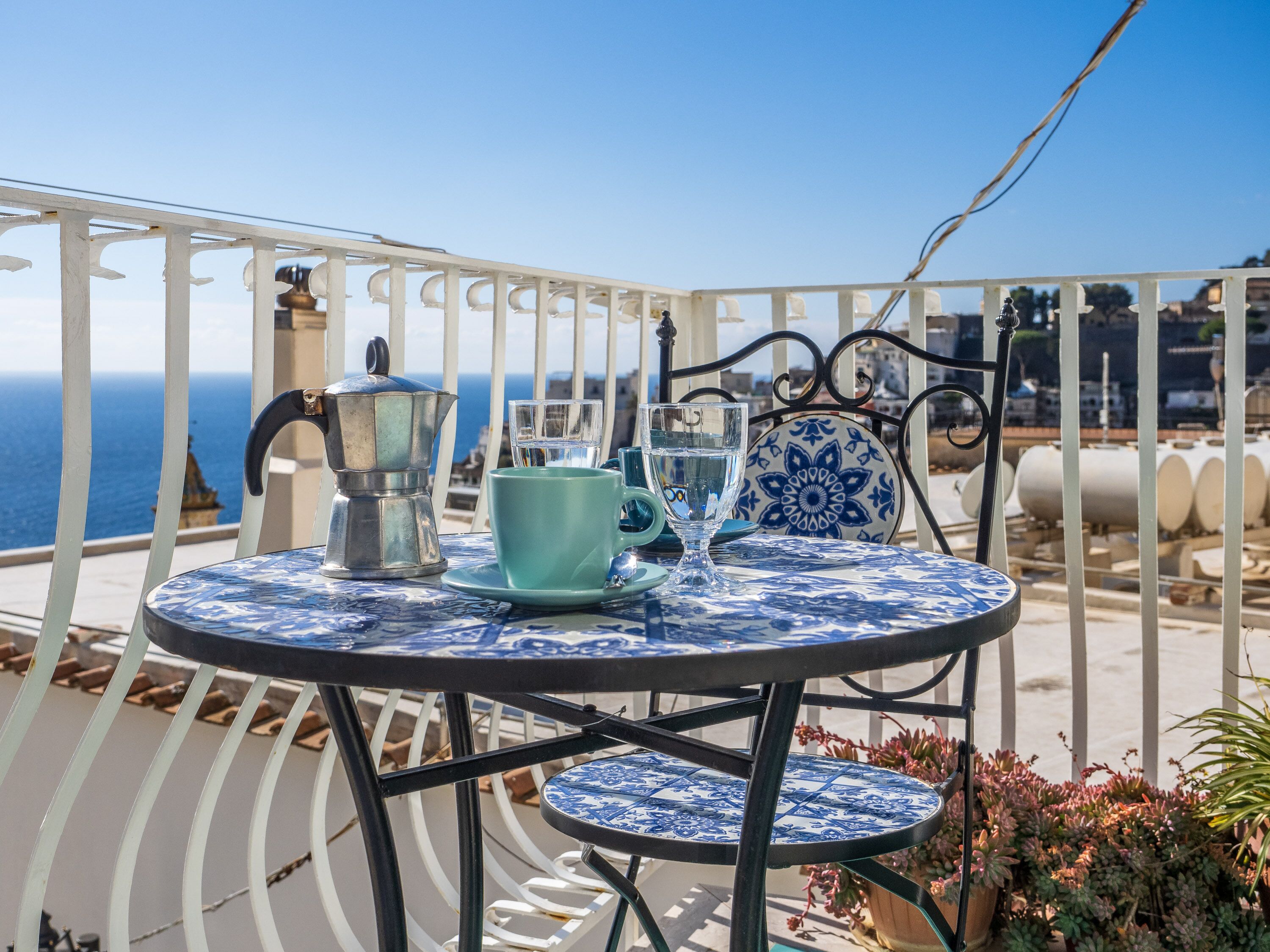 A round table with a blue and white patterned tablecloth, a metal coffee pot, a glass cup, and a matching saucer.
