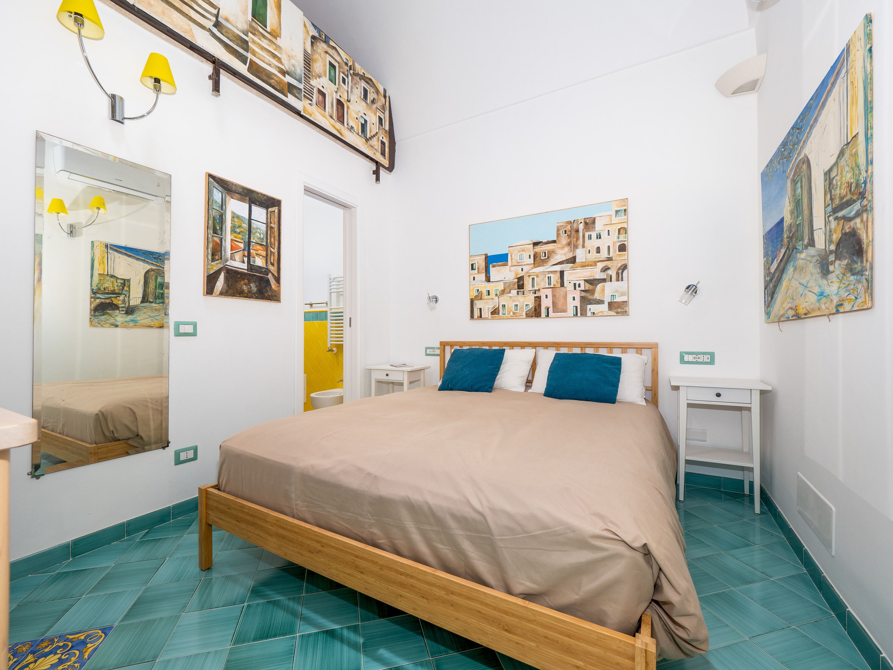 A bedroom with a wooden bed, a nightstand, a bedside table, and a large mirror. There are paintings on the walls and a tiled floor.
